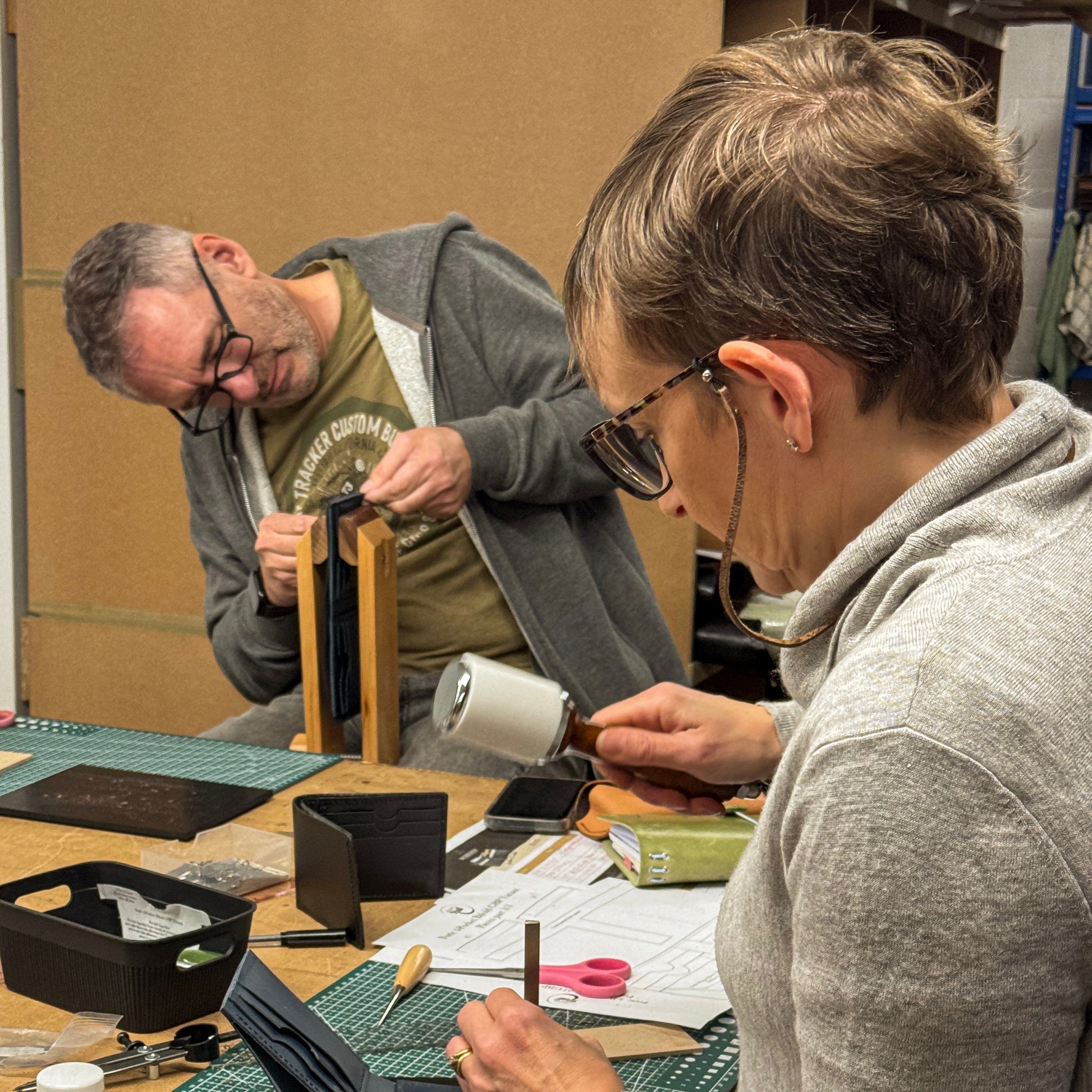 Two people working on a leather project at a table with tools and materials.