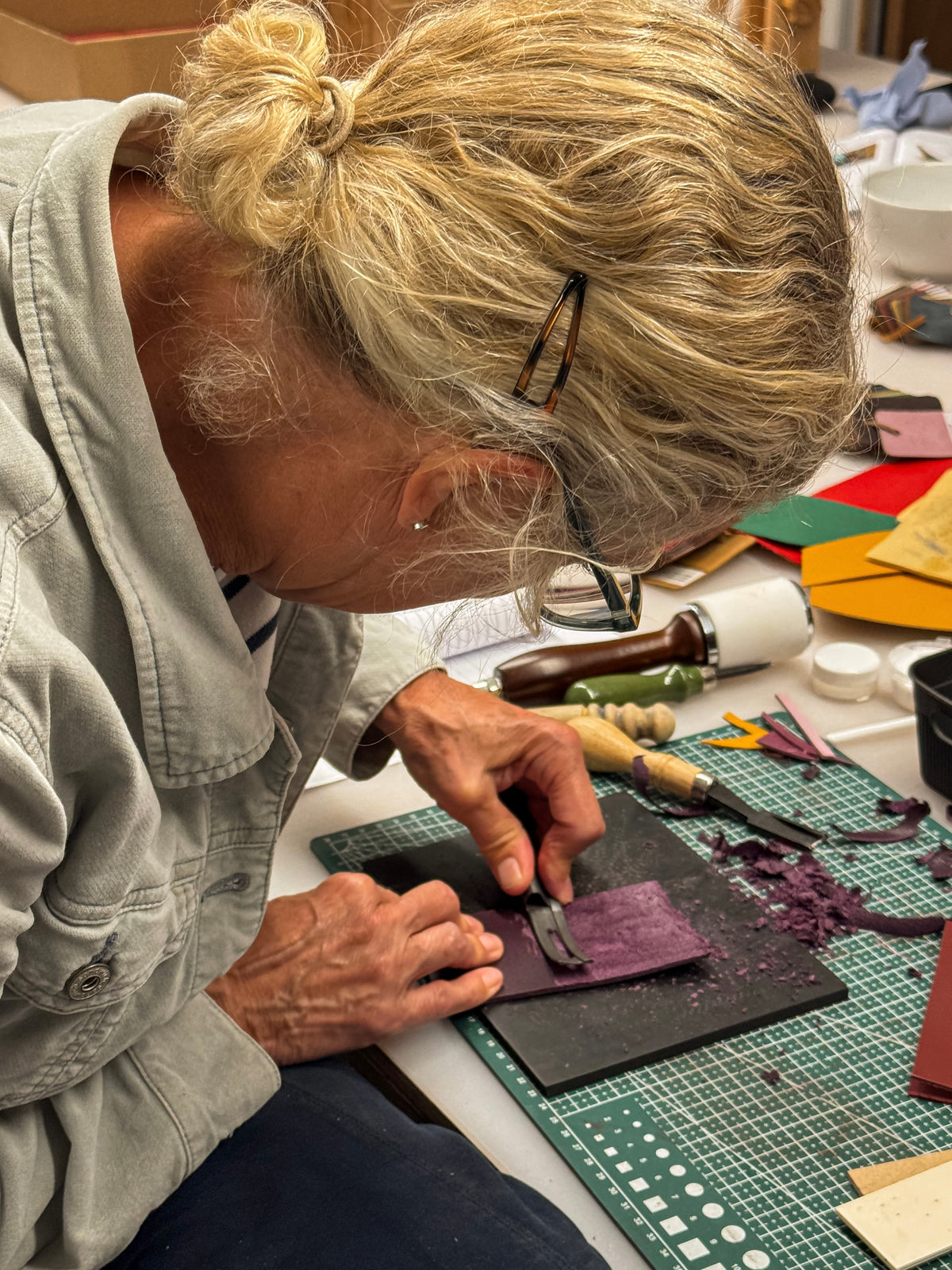 A woman with blonde hair in a bun and glasses is carving a purple rectangular block on a black cutting mat, surrounded by crafting tools and colorful material sheets in a workshop setting.