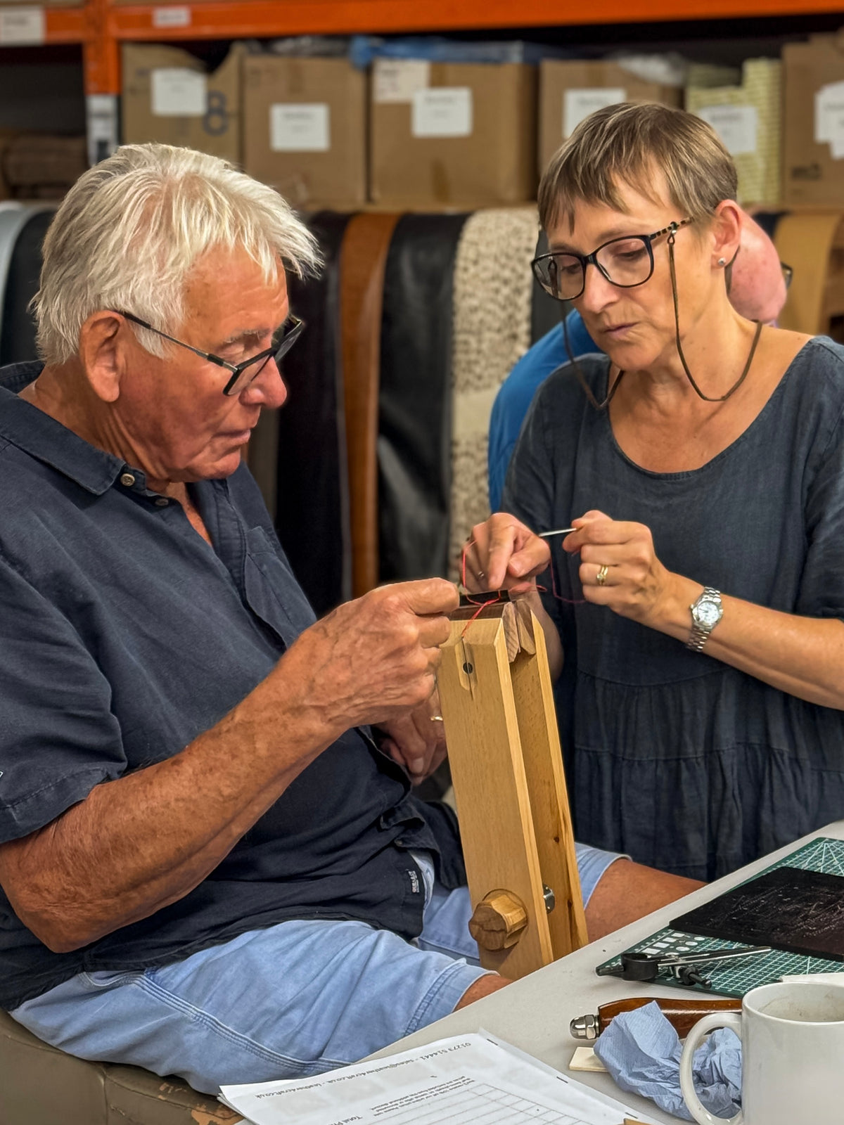 An older man and a woman are seated at a table in a workshop or studio, both wearing glasses and focused on a small wooden object held in the man's hands. Shelves with boxes are visible in the background, along with a mug and papers on the table.