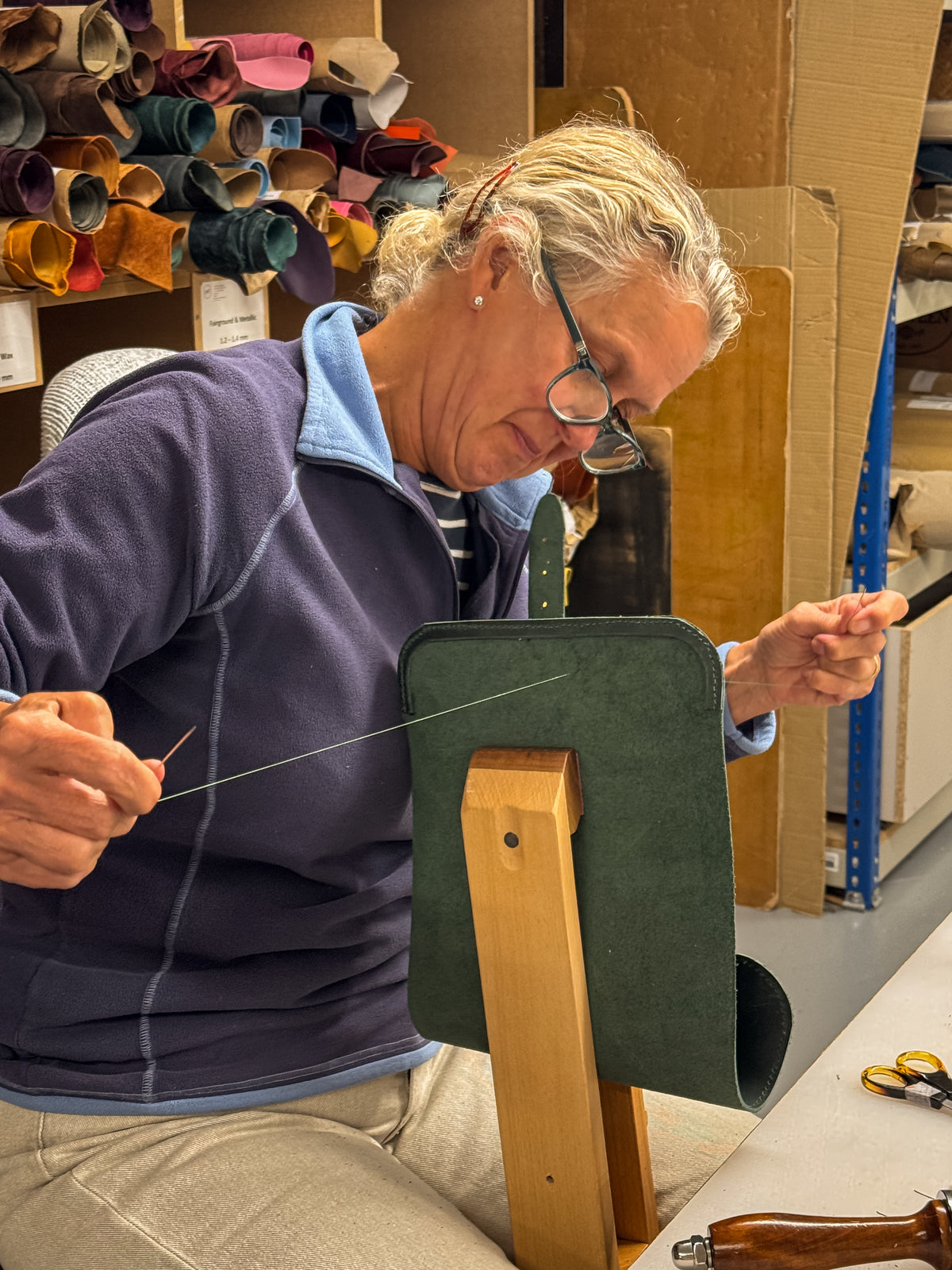 A woman wearing glasses and a navy fleece jacket sits in a leatherworking workshop, holding a leather edge on a wooden stand with a needle in her hand. Colorful rolls of leather are stacked on shelves in the background.