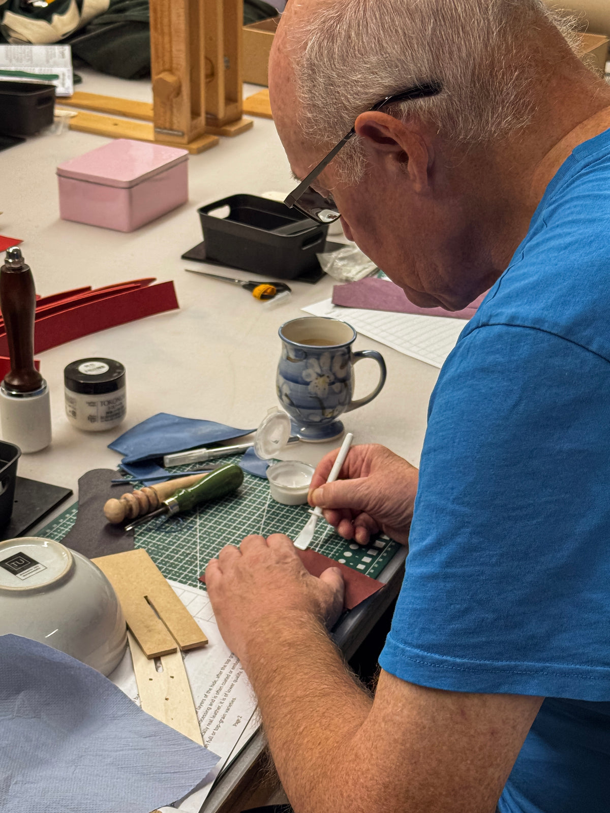 An older man with white hair and glasses, wearing a blue t-shirt, is shown from the side as he works at a cluttered craft table, using a white-handled tool on a dark piece of material. The table is covered with various tools, a blue and white ceramic mug, papers, and small containers.