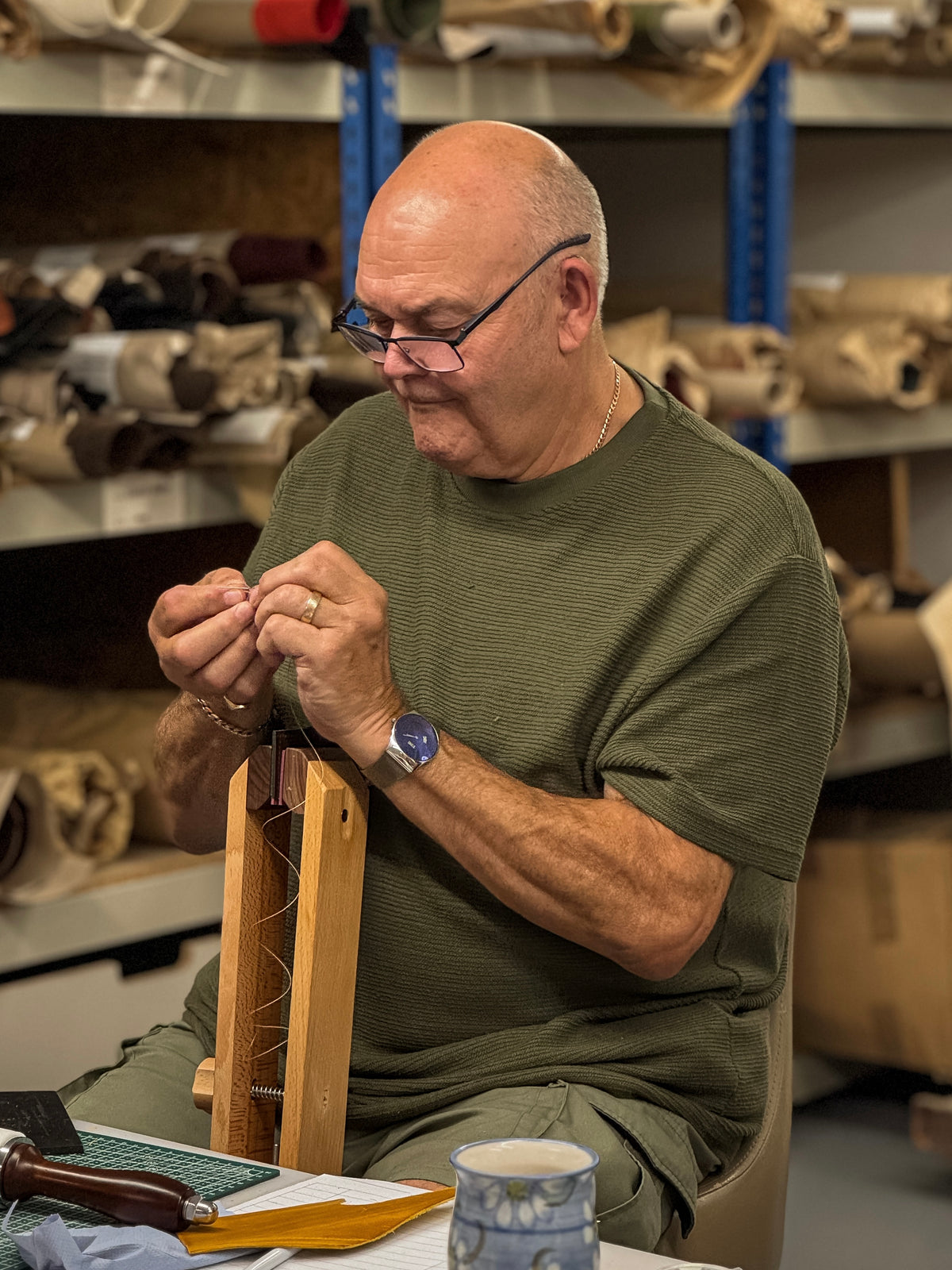A bald man with glasses, wearing an olive green t-shirt, is seated in a workshop, holding a small wooden object in his hands. Shelves filled with fabric rolls are visible in the background.