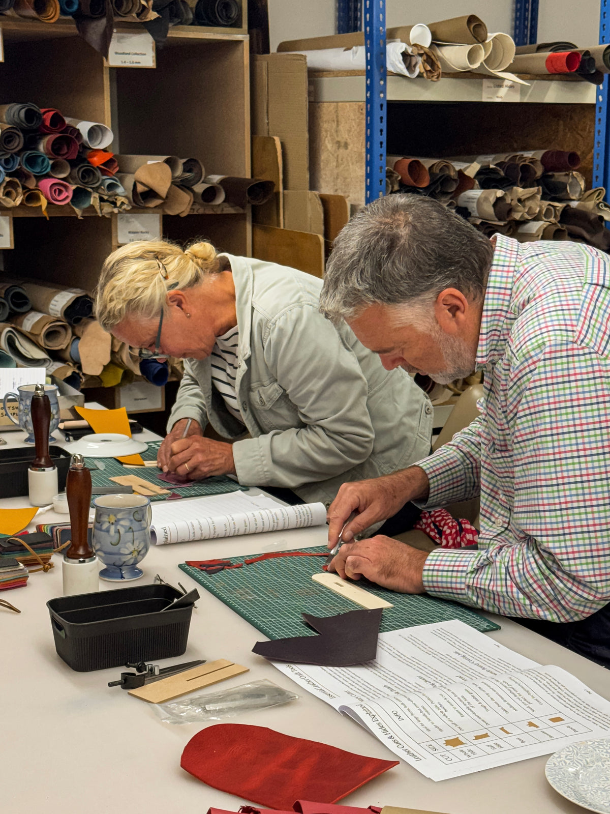 Two artisans working at a table in a leathercraft studio, cutting and preparing leather pieces on a green cutting mat with shelves of leather rolls in the background.