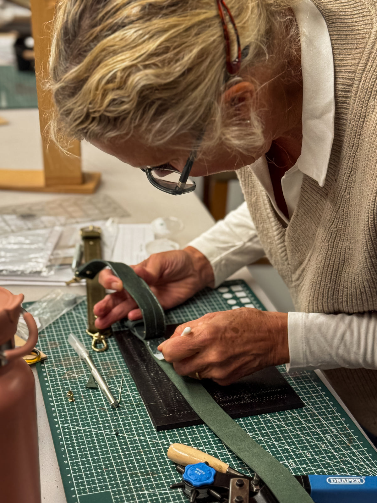 An artisan wearing glasses and a beige sweater is shown crafting a dark green leather strap on a green cutting mat at a worktable.