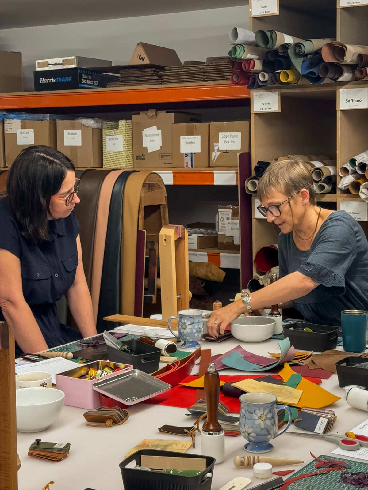 Two women collaborating in a creative studio or workshop, surrounded by craft supplies, leather rolls, and mugs on a cluttered but productive table.