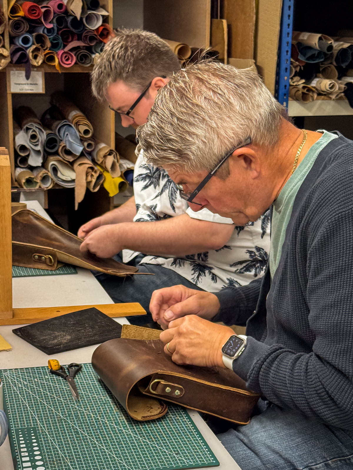 A close-up of a leather craftsman working on a brown leather item at a table, with another person visible in the background among shelves of leather rolls.