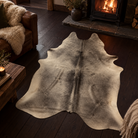 A natural cowhide area rug with a black and cream mottled pattern, displayed on a dark wood floor in a cozy living room setting with a fireplace and leather sofa.