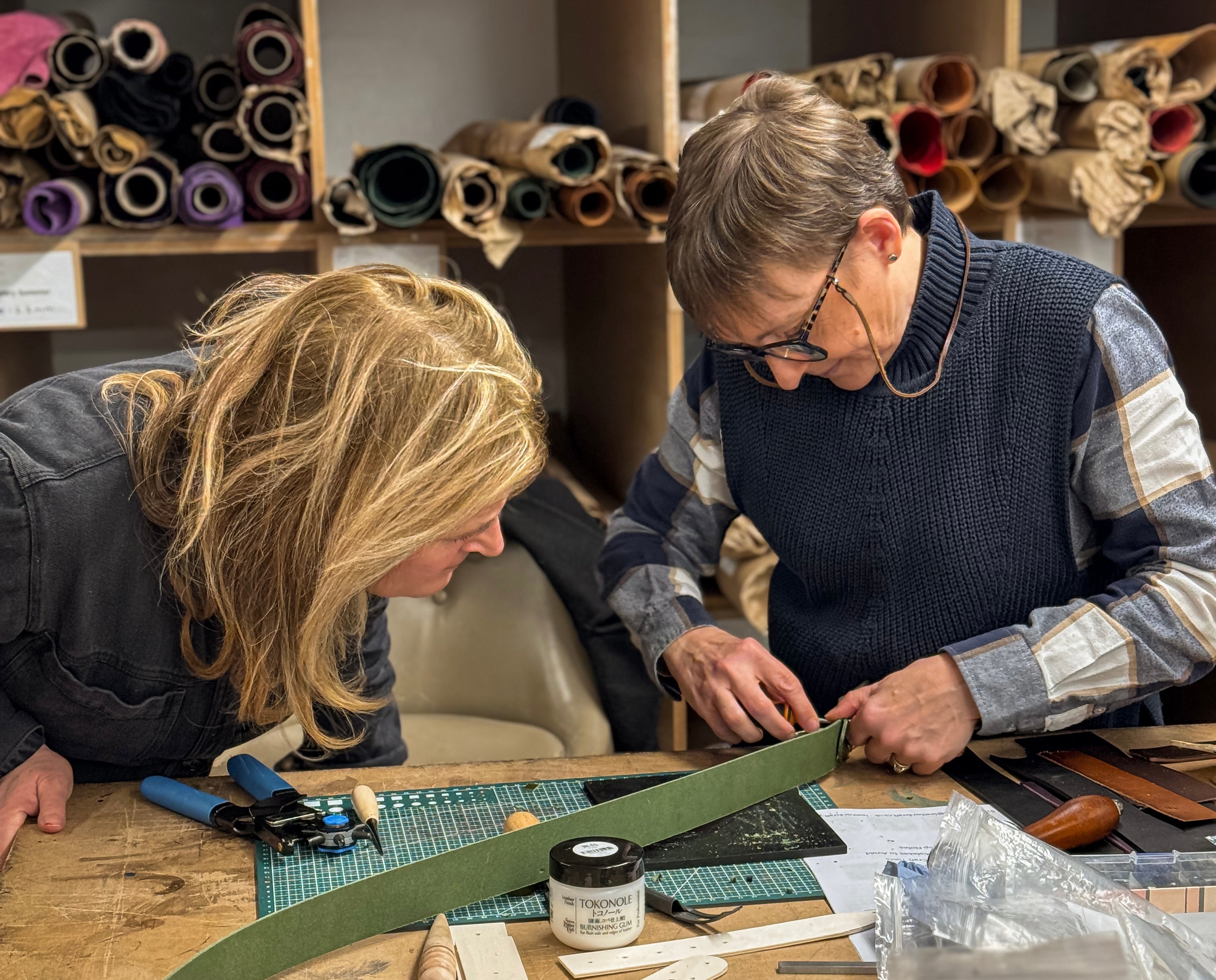 Two women collaborating in a leathercraft workshop, focused on cutting and preparing a long green leather strip on a wooden table surrounded by tools and rolls of material.