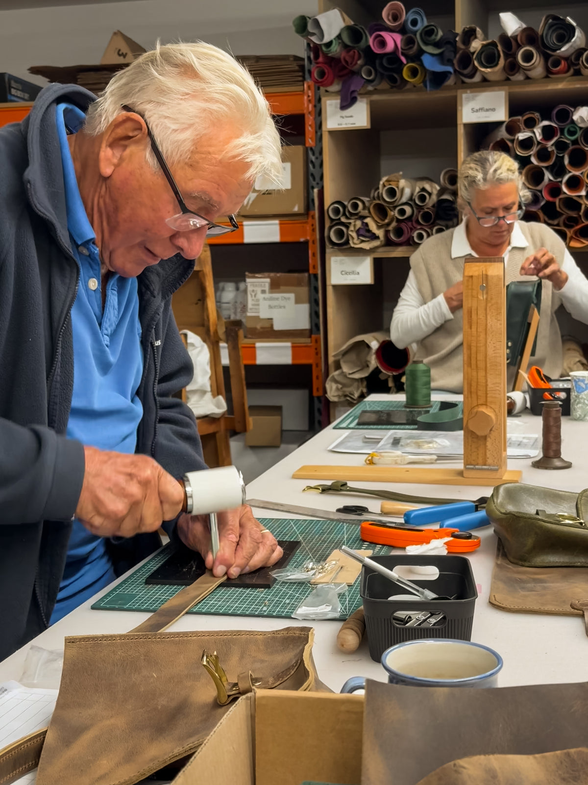 An elderly man with white hair and glasses is shown in a leathercraft workshop, using a leather mallet to strike a piece of leather on a green cutting mat. In the background, another person is working at a table surrounded by leather tools, rolls, and equipment.