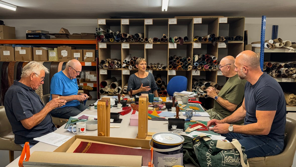 A group of five people seated around a table in a workshop or studio, surrounded by shelves filled with rolled materials. The scene appears to show a hands-on craft or sewing project in progress, with fabric, tools, and work materials on the table.