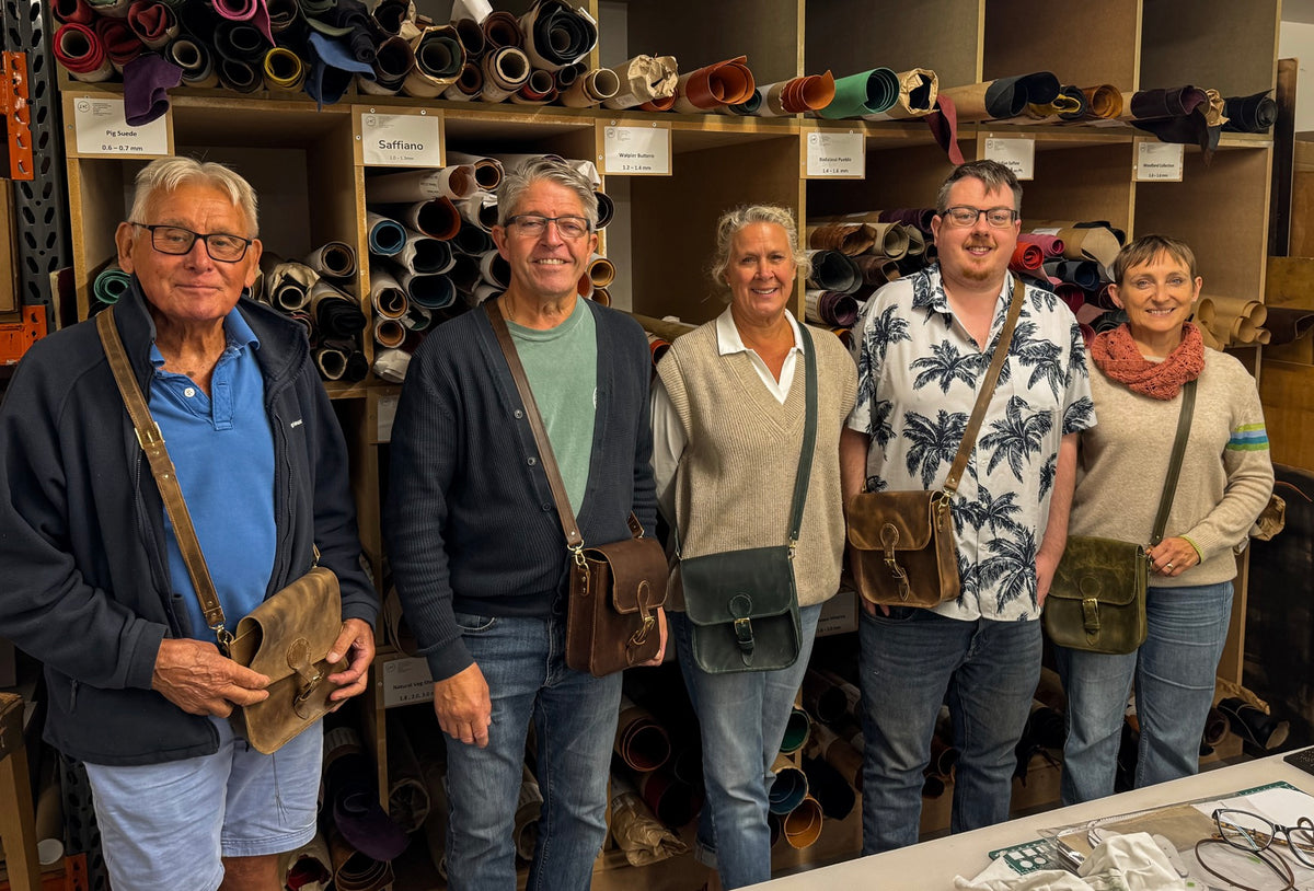 Five people standing together in a leather goods store, each wearing a different leather crossbody or shoulder bag. Shelves behind them are filled with colorful leather rolls.