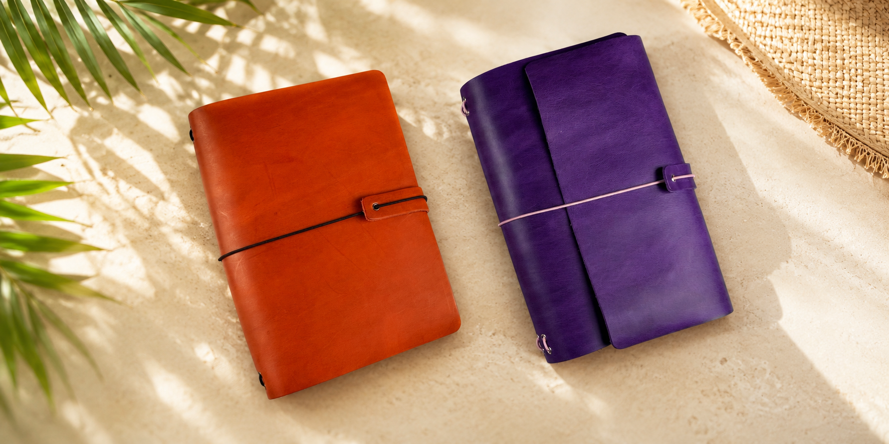Two leather travel notebooks or journals with wraparound straps, one in orange-brown and one in purple, arranged on a light beige surface with palm leaves and a woven mat in the background.