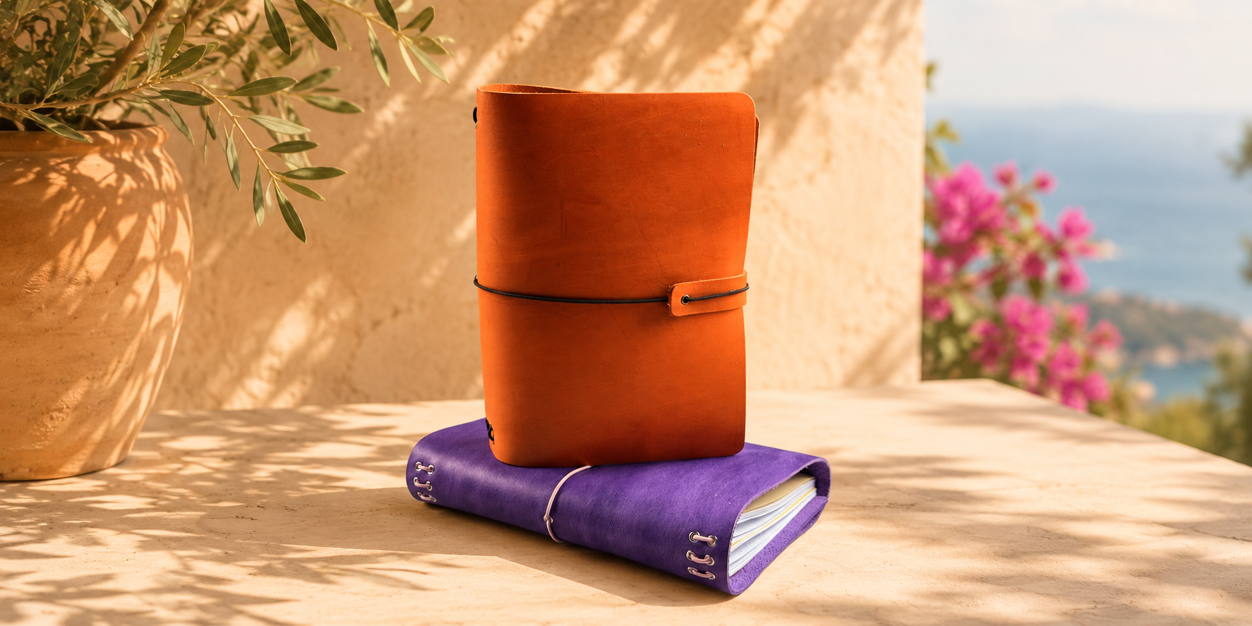 An orange leather journal with a strap closure stacked on top of a purple leather notebook, set on a sunlit stone ledge with a potted plant and outdoor scenery in the background.