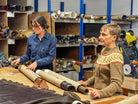 Two women in a warehouse setting, one rolling leather and the other holding it, with shelves and equipment in the background.