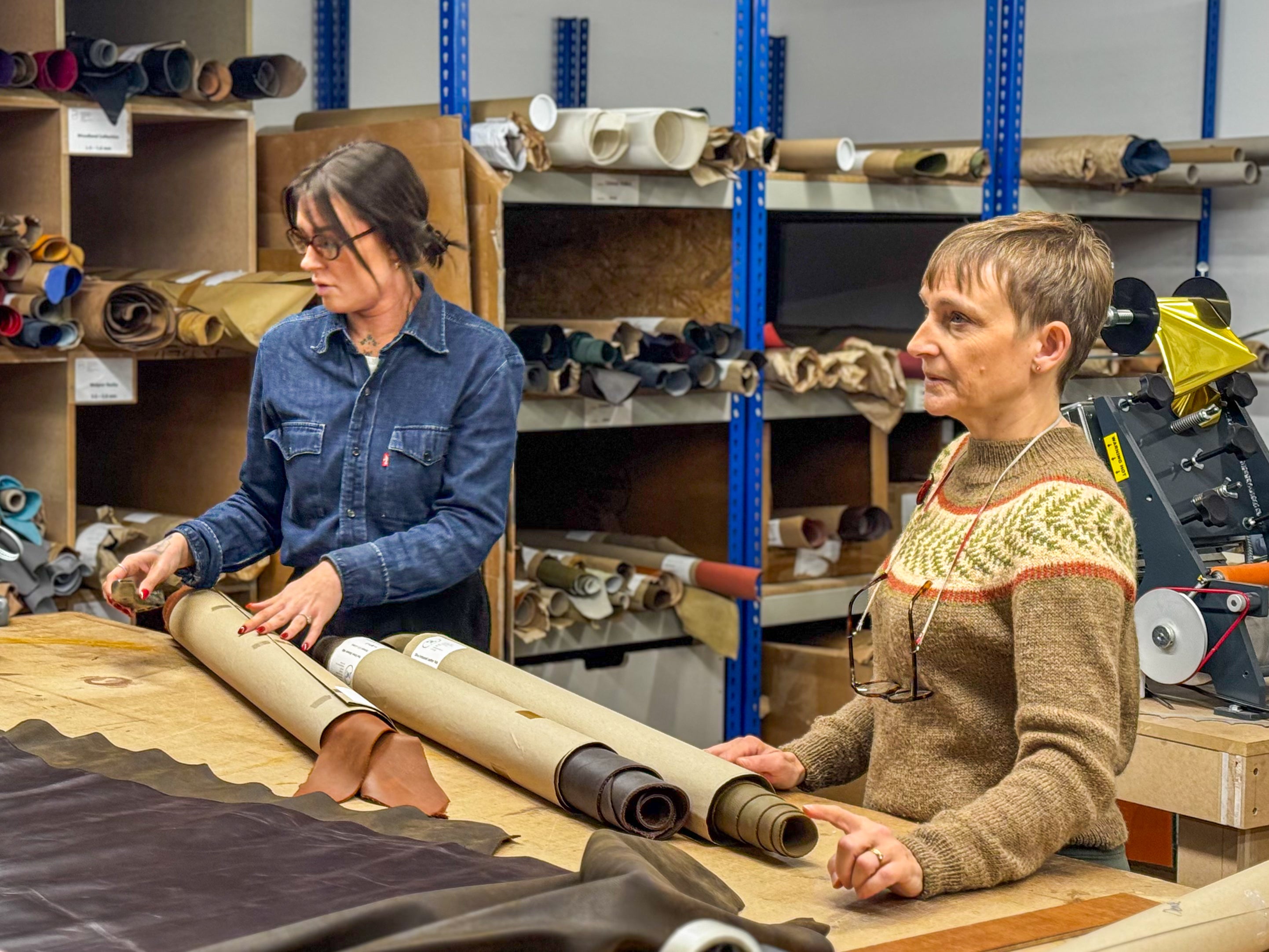 Two women in a warehouse setting, one rolling leather and the other holding it, with shelves and equipment in the background.