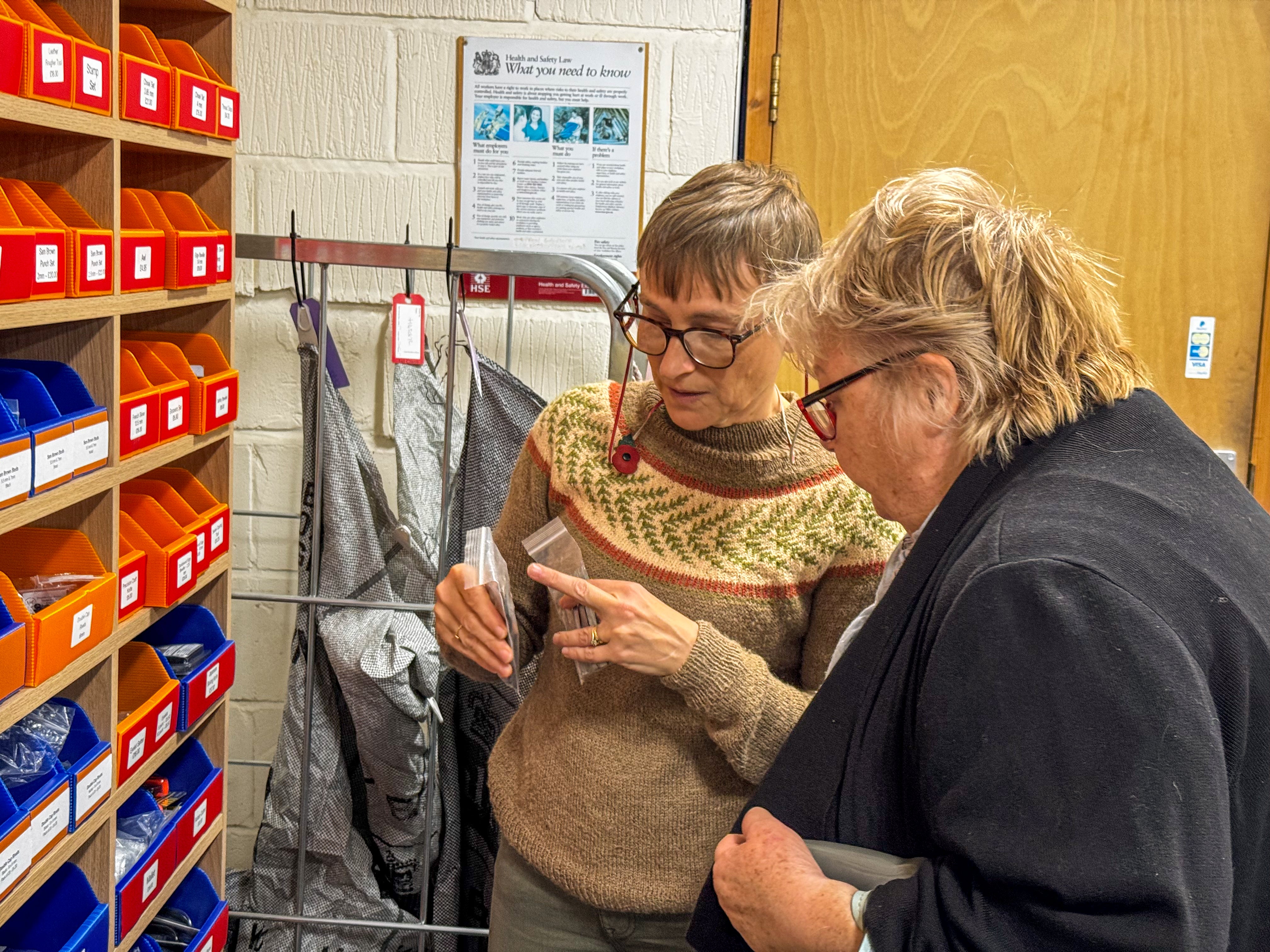Two people looking at a leather tools in a workshop with shelves and posters in the background.