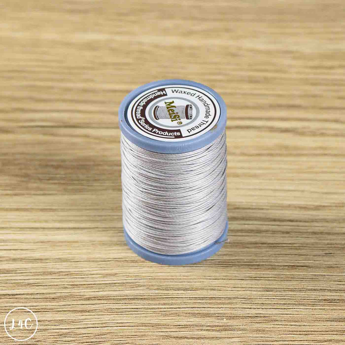 A spool of Melody Waxed Merino Wool Thread in a light gray color, resting on a light wood grain surface. 