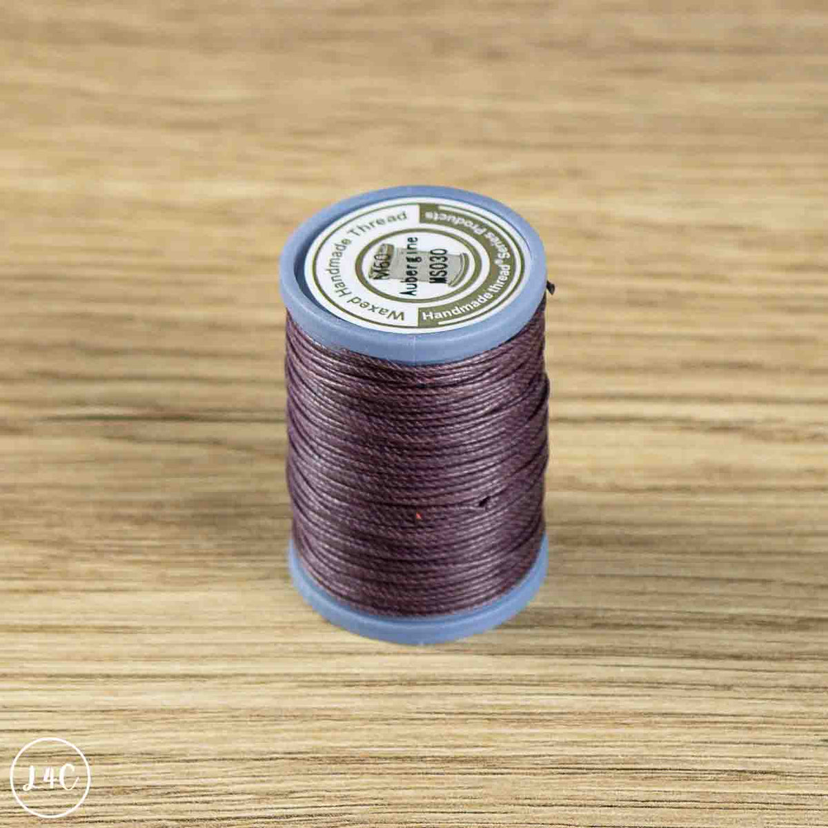 A spool of sewing thread in a dark plum purple color, standing upright on a light brown wooden surface. The spool has a blue plastic rim and a white label on top.