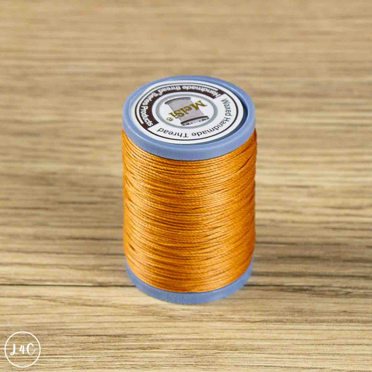 A spool of MAS-14C embroidery thread in a warm golden-orange color, wound on a blue plastic core and placed on a light wood grain surface.