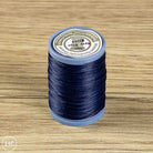 A spool of sewing thread in the color Navy Blue, placed on a light brown wooden surface. The spool has a blue plastic base and a white paper label on top.