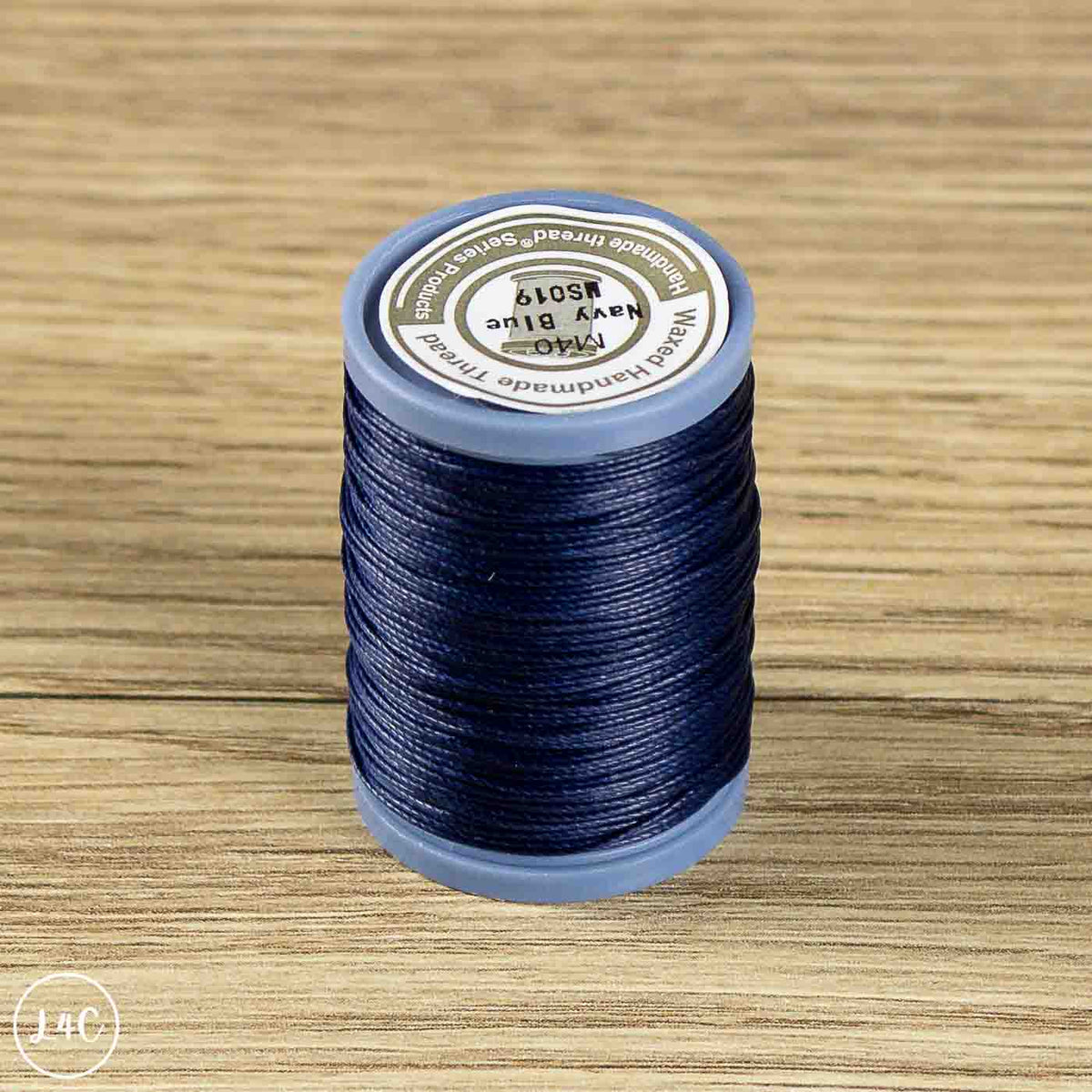 A spool of sewing thread in the color Navy Blue, placed on a light brown wooden surface. The spool has a blue plastic base and a white paper label on top.