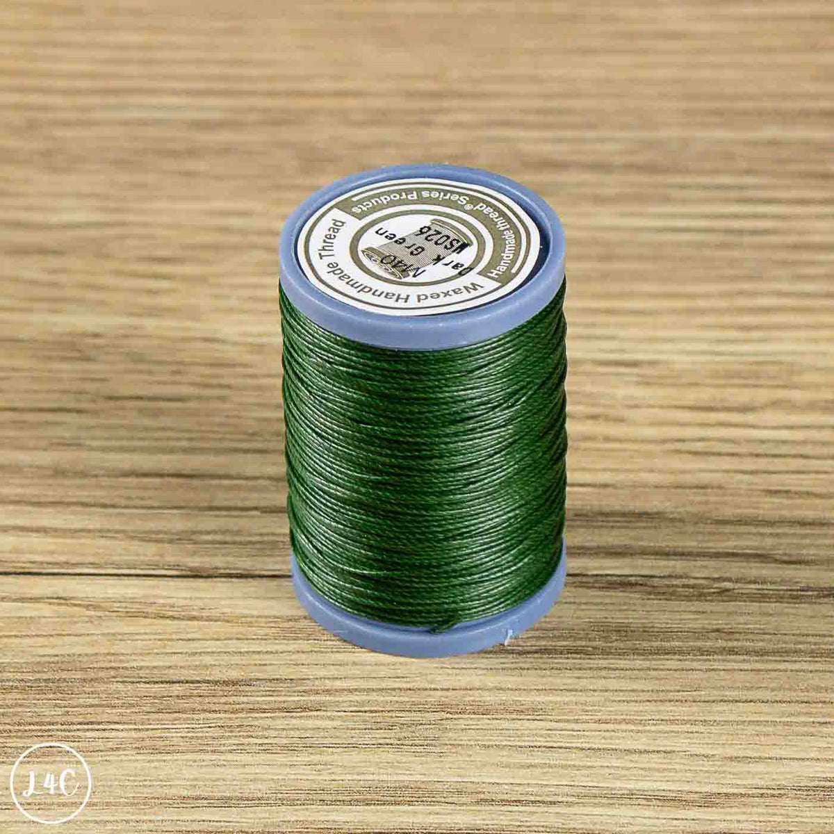 A spool of green sewing thread standing upright on a light wood-grain surface. The spool has a blue plastic base and a white circular label on top with black text.