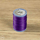A spool of purple sewing thread standing upright on a light brown wood-grain surface. The spool has a light blue plastic rim and a white paper label.