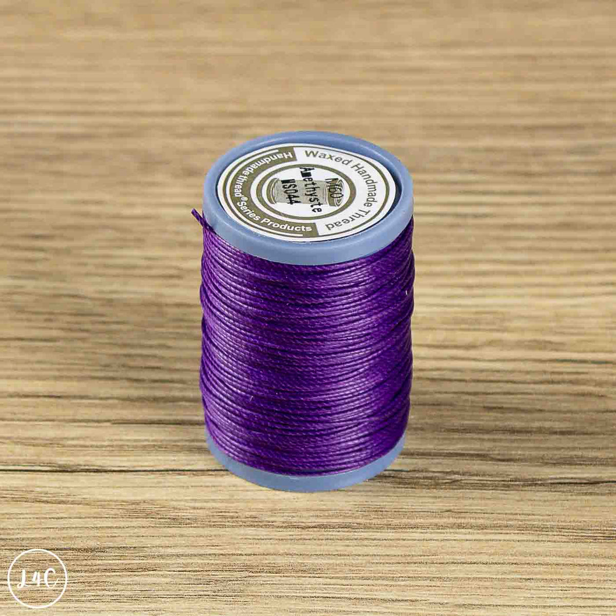 A spool of purple sewing thread standing upright on a light brown wood-grain surface. The spool has a light blue plastic rim and a white paper label.