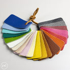 A leather color swatch fan showing multiple textured leather color options in blue, green, white, beige, pink, yellow, brown, orange, and red, tied together with a brown leather loop on a light gray background.