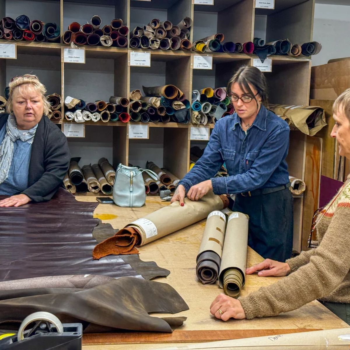 Three women in a workshop surrounded by leather rolls and sheets on a worktable, with shelves of leather material rolls in the background.